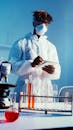 A Man Wearing Face Mask and Lab Coat While Standing by the Table With His Experiments