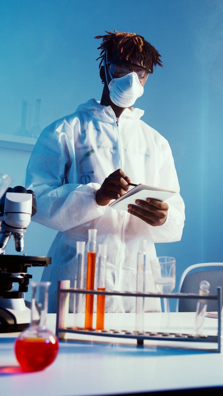 A Man Wearing Face Mask And Lab Coat While Standing By The Table With His Experiments