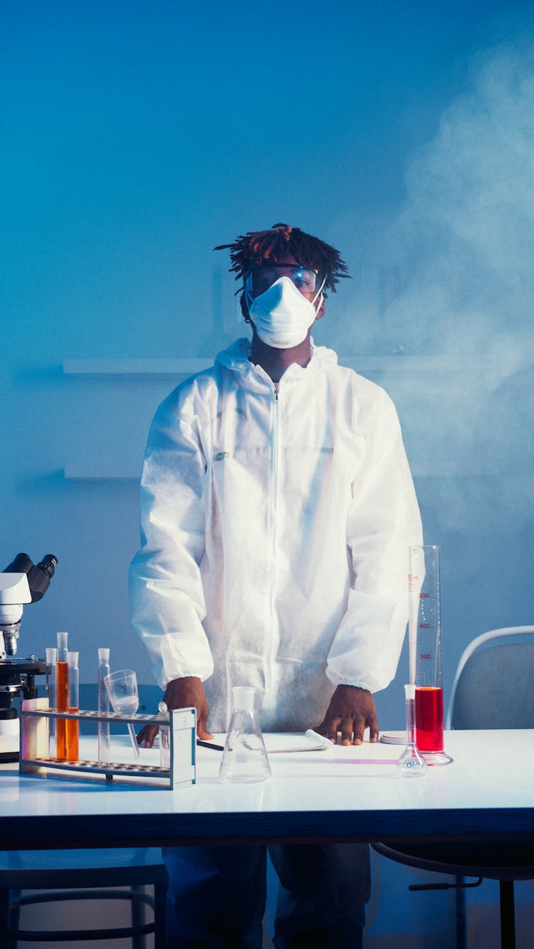 A Man Wearing Face Mask And Lab Coat While Standing By The Table With His Experiments