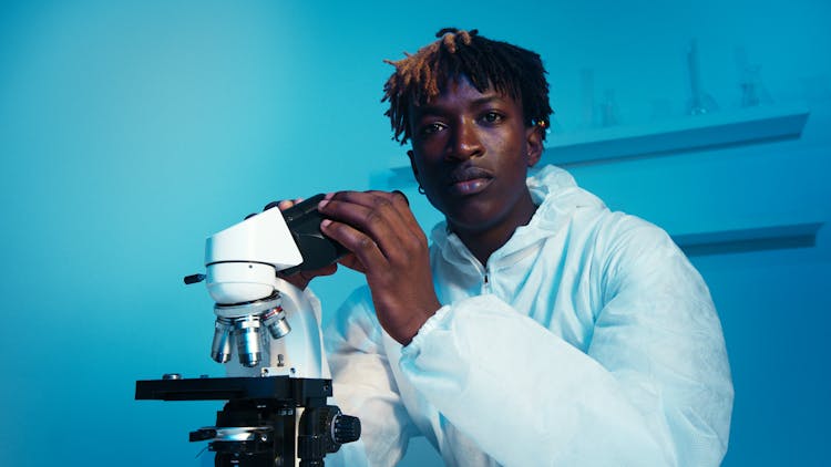 A Man Looking At The Camera While Holding The Eyepiece Of The Microscope