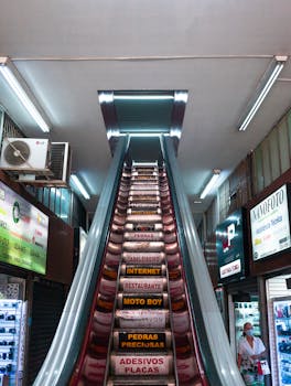 Vertical view of a brightly lit indoor escalator in a shopping center with various store signs.