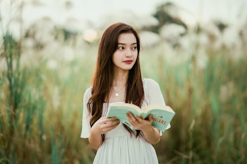 Young contemplative ethnic female in pendant with open textbook looking away in countryside on summer day