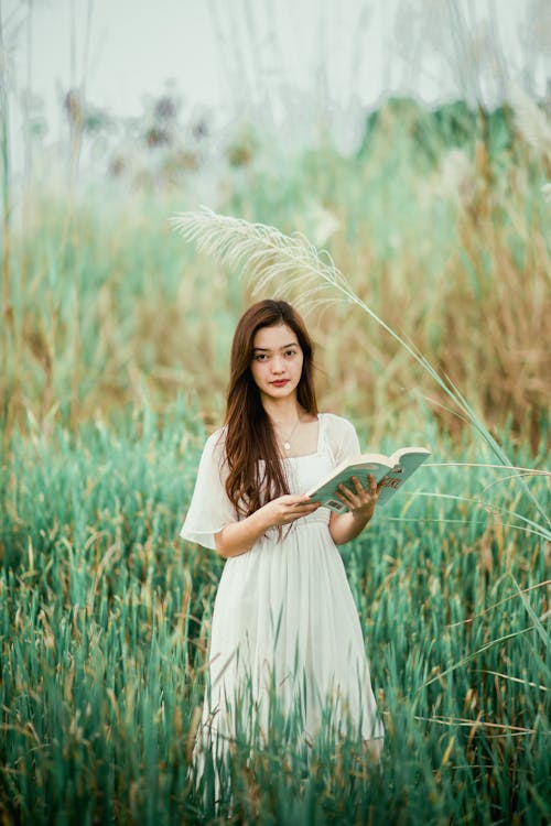 Free Young charming Asian female in dress with open book looking at camera on green meadow Stock Photo