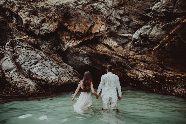 Anonymous Young Couple Holding Hand And Walking In Sea Water Near Rocky Grotto