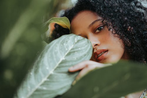 Beautiful serene ethnic female with black curly hair standing in garden and touching green plant leaves while looking at camera