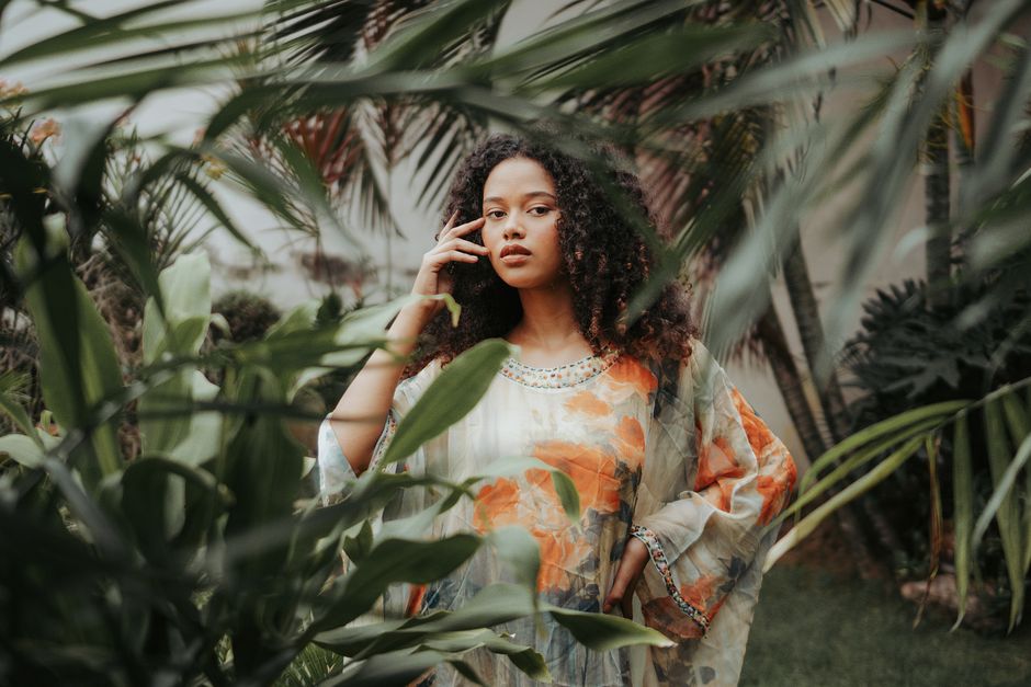Elegant pretty ethnic female in trendy loose dress touching face gracefully and looking at camera while standing with arm on waist in green tropical garden
