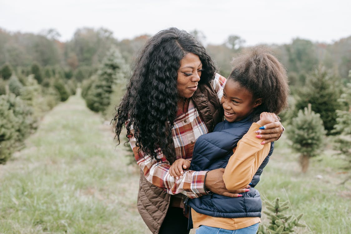 Free Cheerful African American mum cuddling child while standing close among rows of Christmas trees Stock Photo