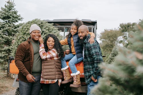 Cheerful African American parents embracing daughter and son near transport and coniferous trees while looking at camera in countryside