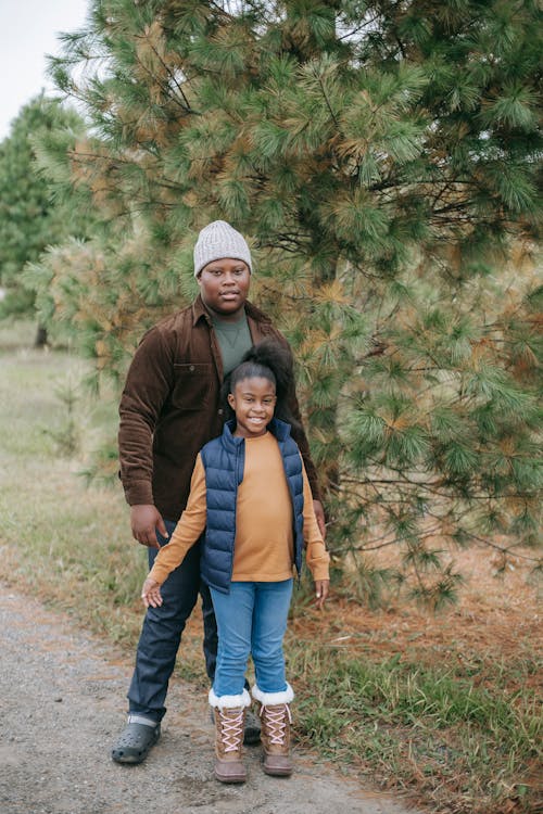 African American teenager with glad sister in casual wear looking at camera on road near fir trees