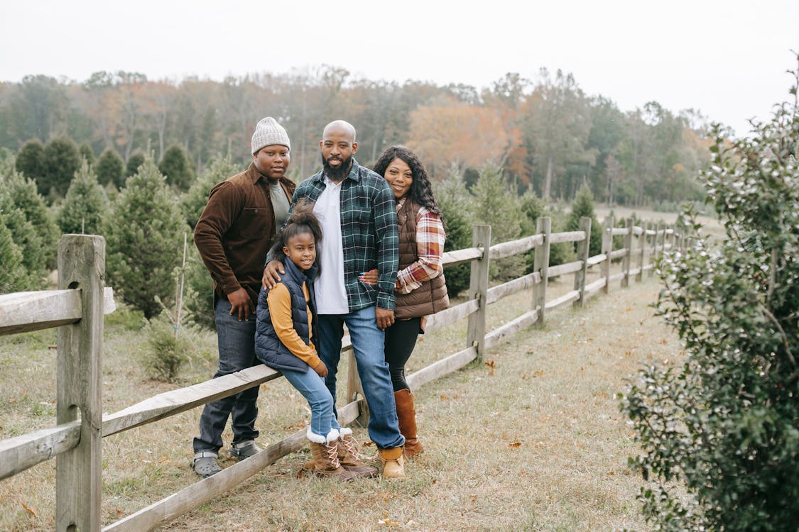 Young content African American parents embracing daughter and son near wooden fence on tree farm while looking at camera
