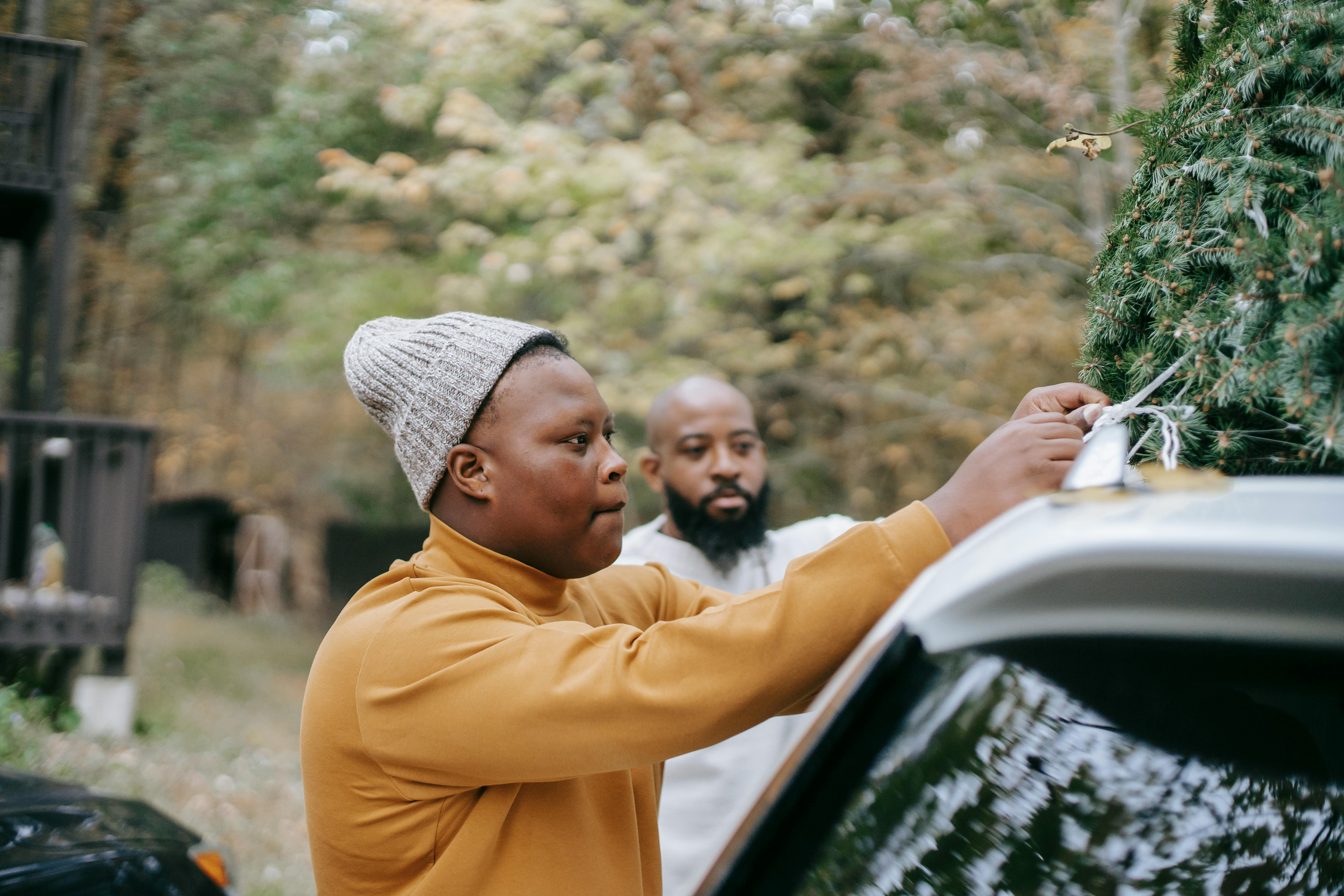 black teen tying fir tree on car roof near father