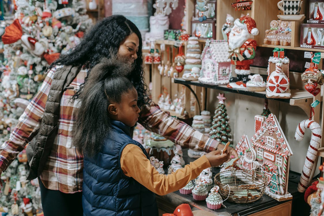 Free Young smiling ethnic woman with girl choosing gifts near Christmas tree in souvenir shop Stock Photo consumers deals