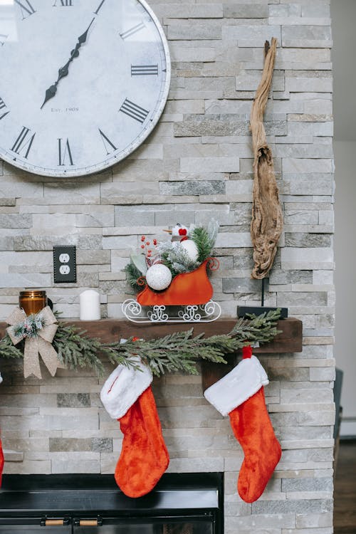 Round clock on wall over shelf with decorative Christmas socks and bright sleigh in house