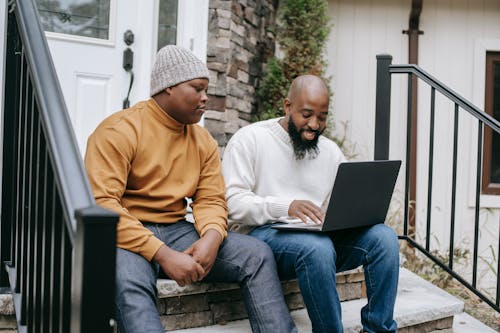 Positive bearded African American male remote worker sitting on stairs and typing on netbook keyboard with teen son