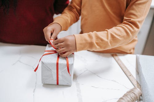 Crop ethnic child tying ribbon on gift box at home