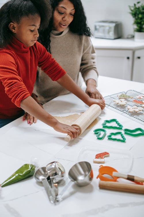 Crop focused African American child rolling out dough near parent while preparing gingerbread cookies at home