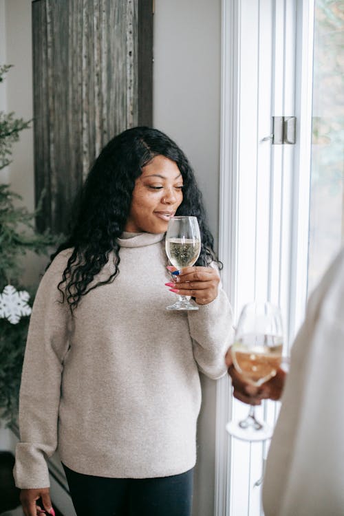 Young content African American female with glass of alcoholic drink speaking with unrecognizable friend while celebrating Christmas at home