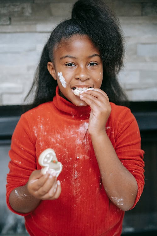 Free Sincere African American child with flour on face tasting crunchy Christmas biscuit while looking at camera at home Stock Photo