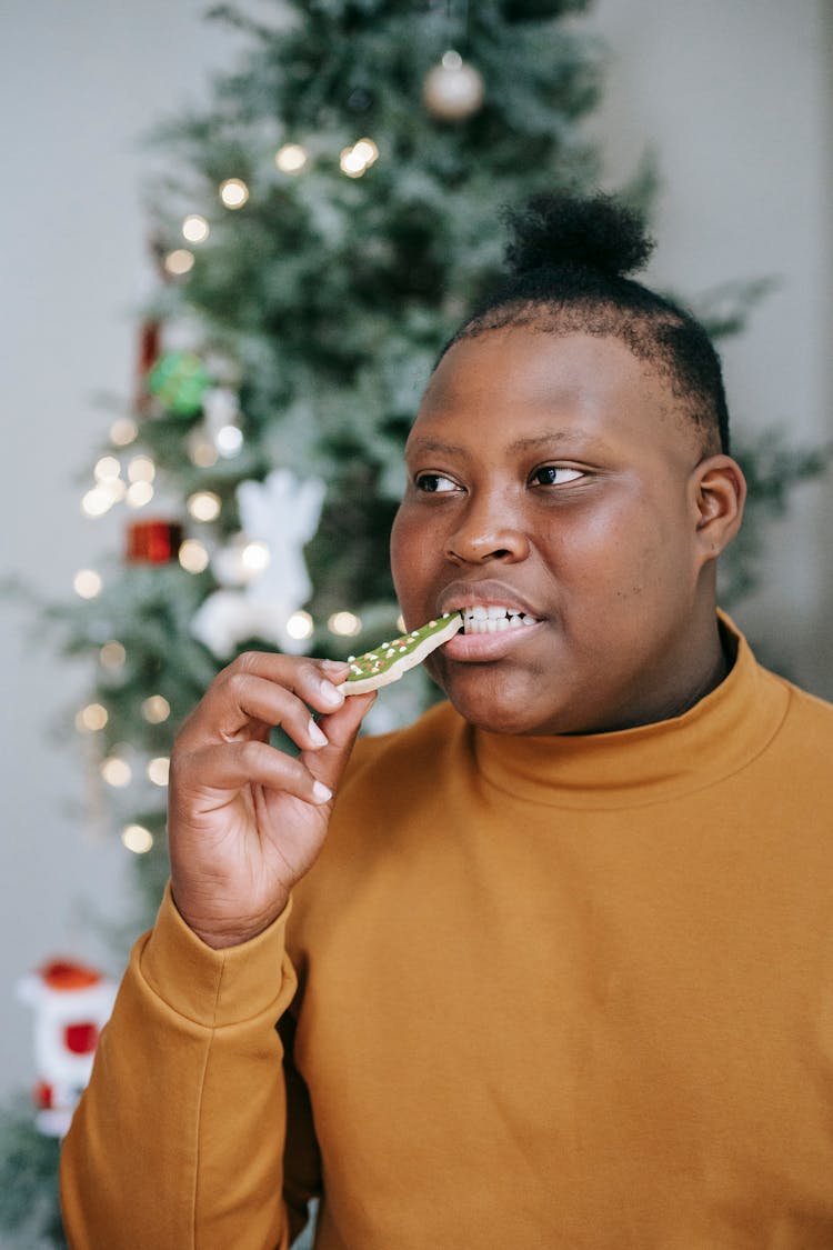 Black Teen Eating Christmas Biscuit Near Fir Tree At Home