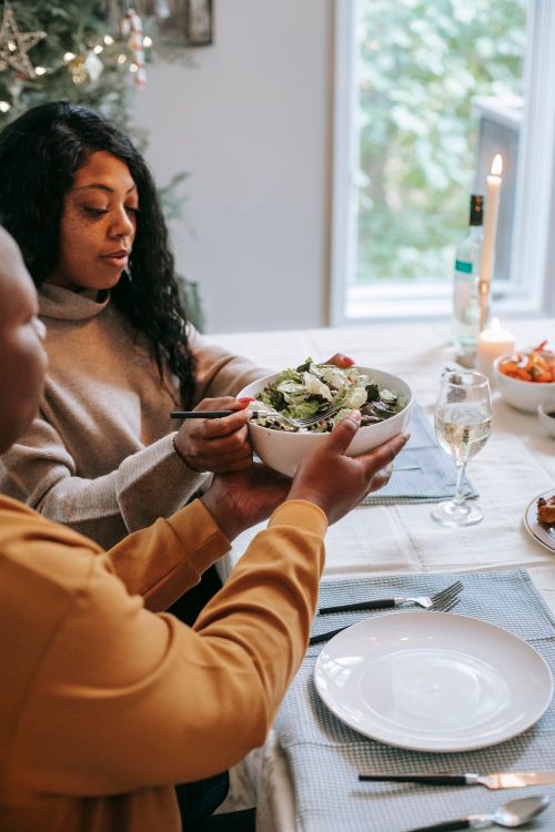 Free Ethnic woman passing vegetable salad to unrecognizable teenager while sitting at table and celebrating New Year holiday in house Stock Photo