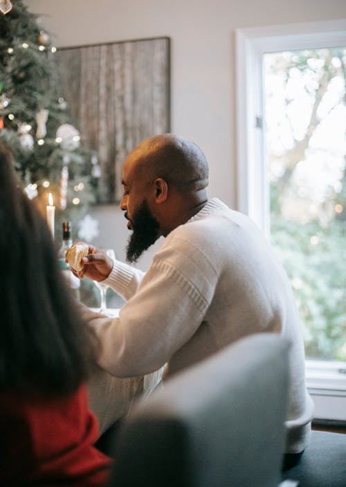 Side view of bearded African American male sitting at table while having dinner during Christmas celebration at home with fir