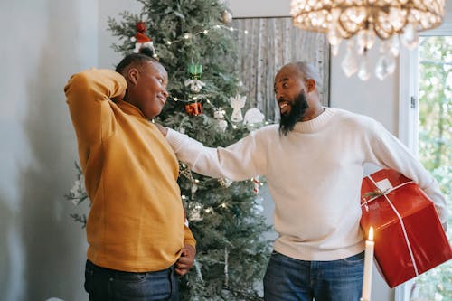 Happy bearded African American man with red box touching shoulder of cheerful black son with hand behind head during Christmas holiday while looking at each other