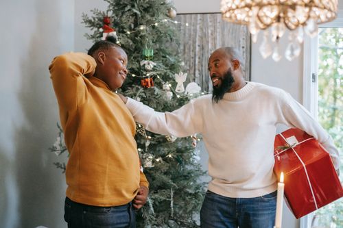 Free Happy bearded African American man with red box touching shoulder of cheerful black son with hand behind head during Christmas holiday while looking at each other Stock Photo