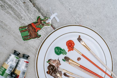 Flat lay of Christmas deer near tubes placed near plate with brushes and mixture of colorful paints