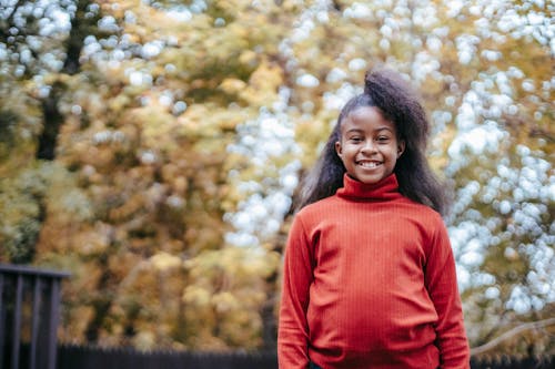 Adorable small African American cheerful girl in pullover looking at camera and smiling on blurred background of autumn trees