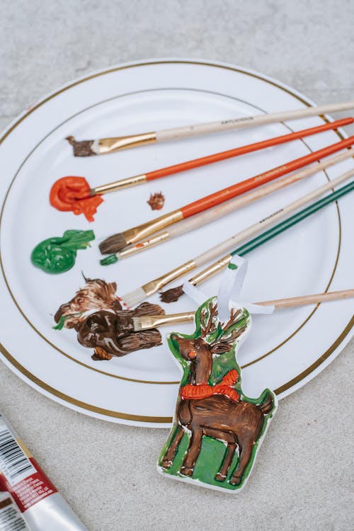 From above of different brushes near mixture of bright paint and colorful small Christmas reindeer figurine