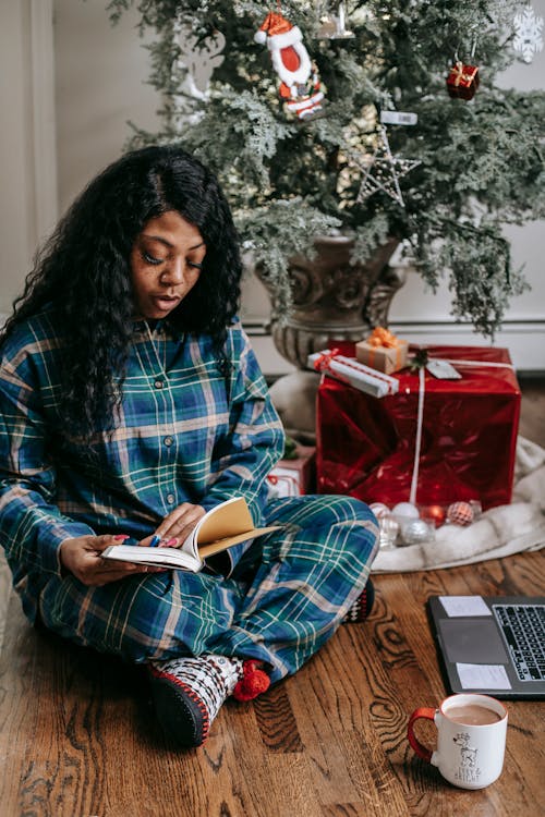 Focused black woman reading book under Christmas tree