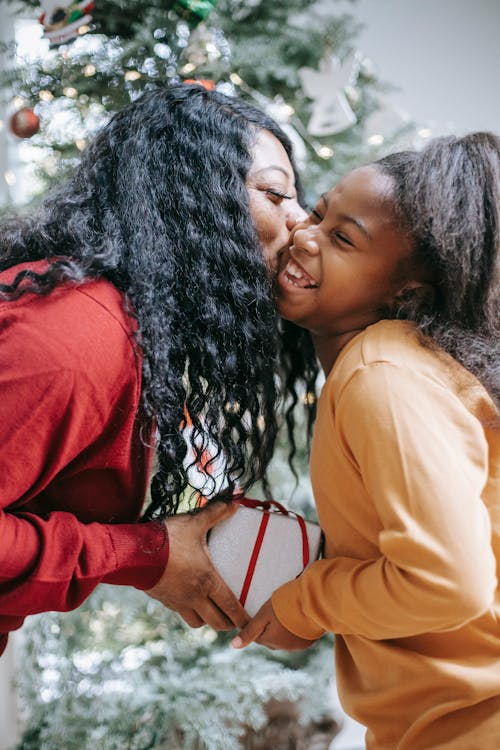 Side view of smiling African American mum kissing cheerful daughter while giving Christmas gift box