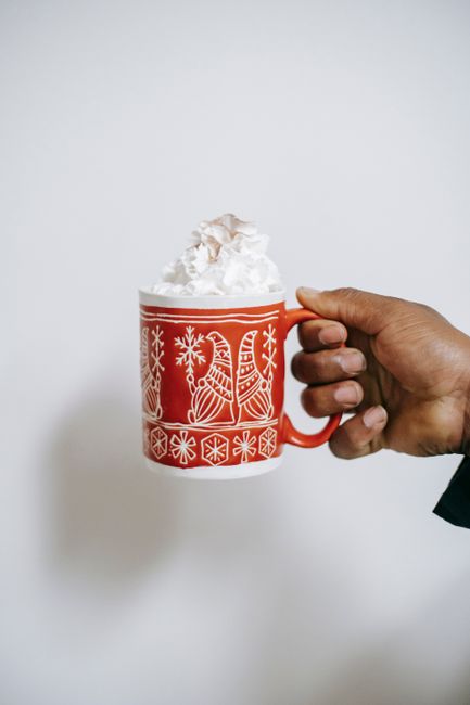 A hand holds a red festive mug filled with a creamy drink topped with whipped cream.