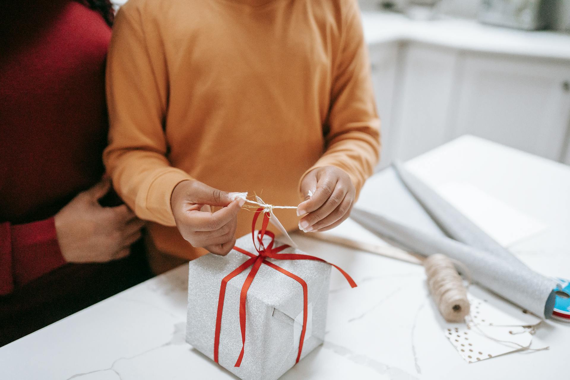 Child wrapping a festive Christmas gift with red ribbon, assisted by an adult, on a white table.