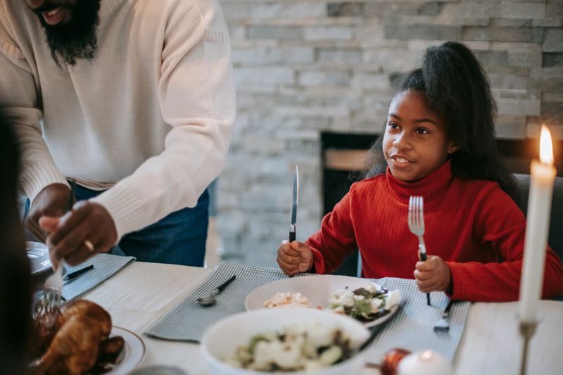 Smiling African American girl sitting at table near father cutting meat during festive dinner with burning candles