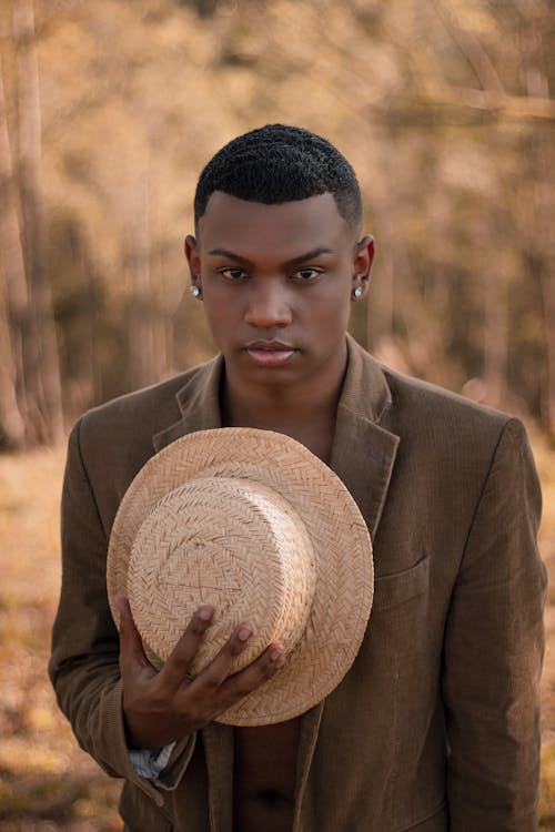 Young African American male in stylish jacket with earrings and straw hat in hands looking at camera