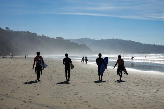 A group of surfers walking along the San Diego beach with surfboards on a sunny day.