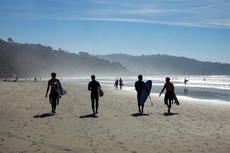 A group of surfers walking along the San Diego beach with surfboards on a sunny day.