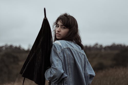 Unemotional young female in denim jacket with closed black umbrella looking at camera over shoulder