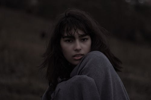 Unemotional young female cuddling in soft plaid looking at camera while standing on hill