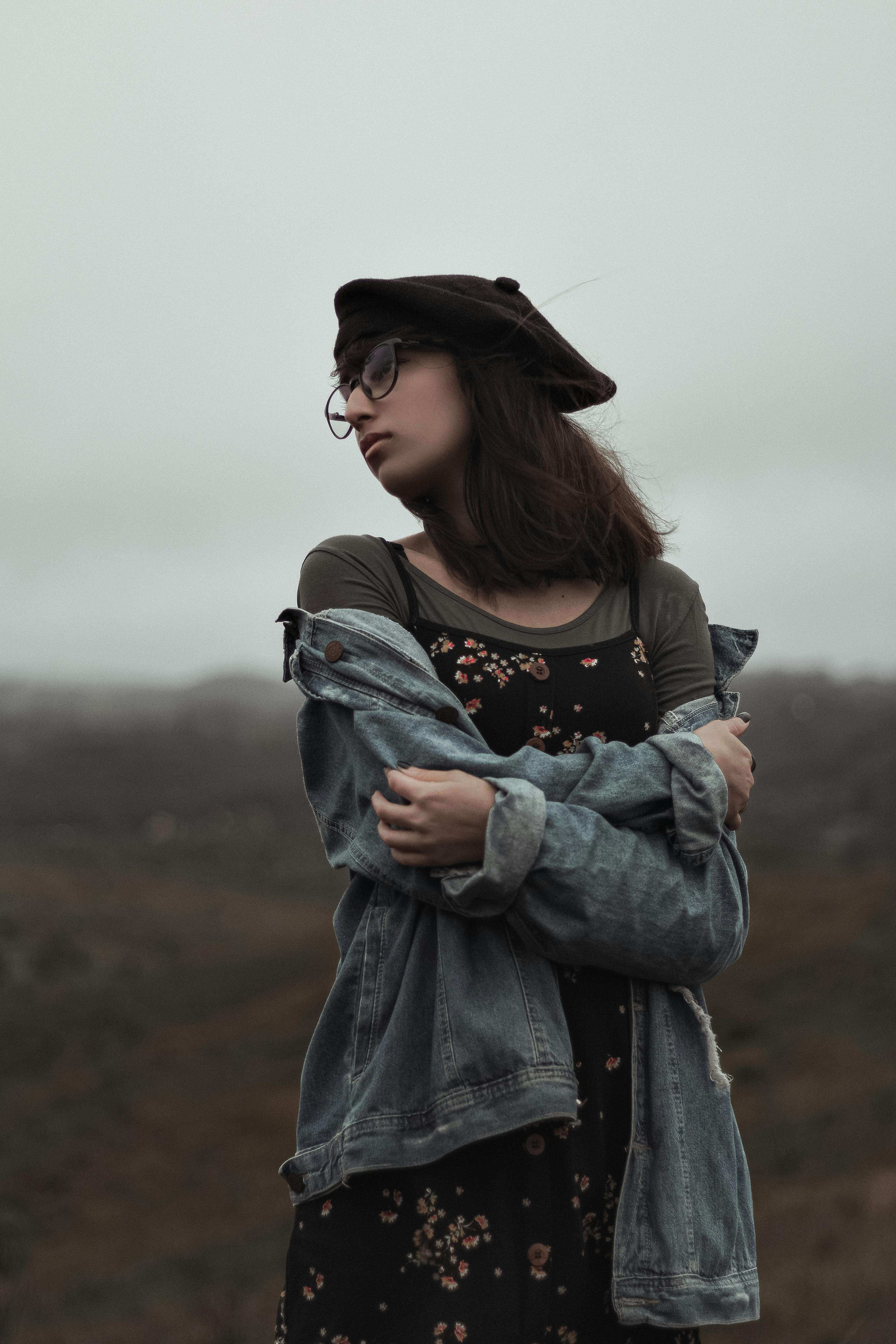 Lonely female in denim jacket and hat with glasses standing in field with arms folded and looking away thoughtfully