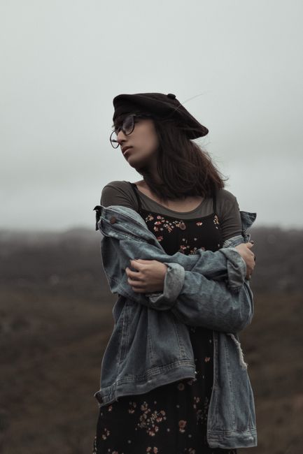 Lonely female in denim jacket and hat with glasses standing in field with arms folded and looking away thoughtfully