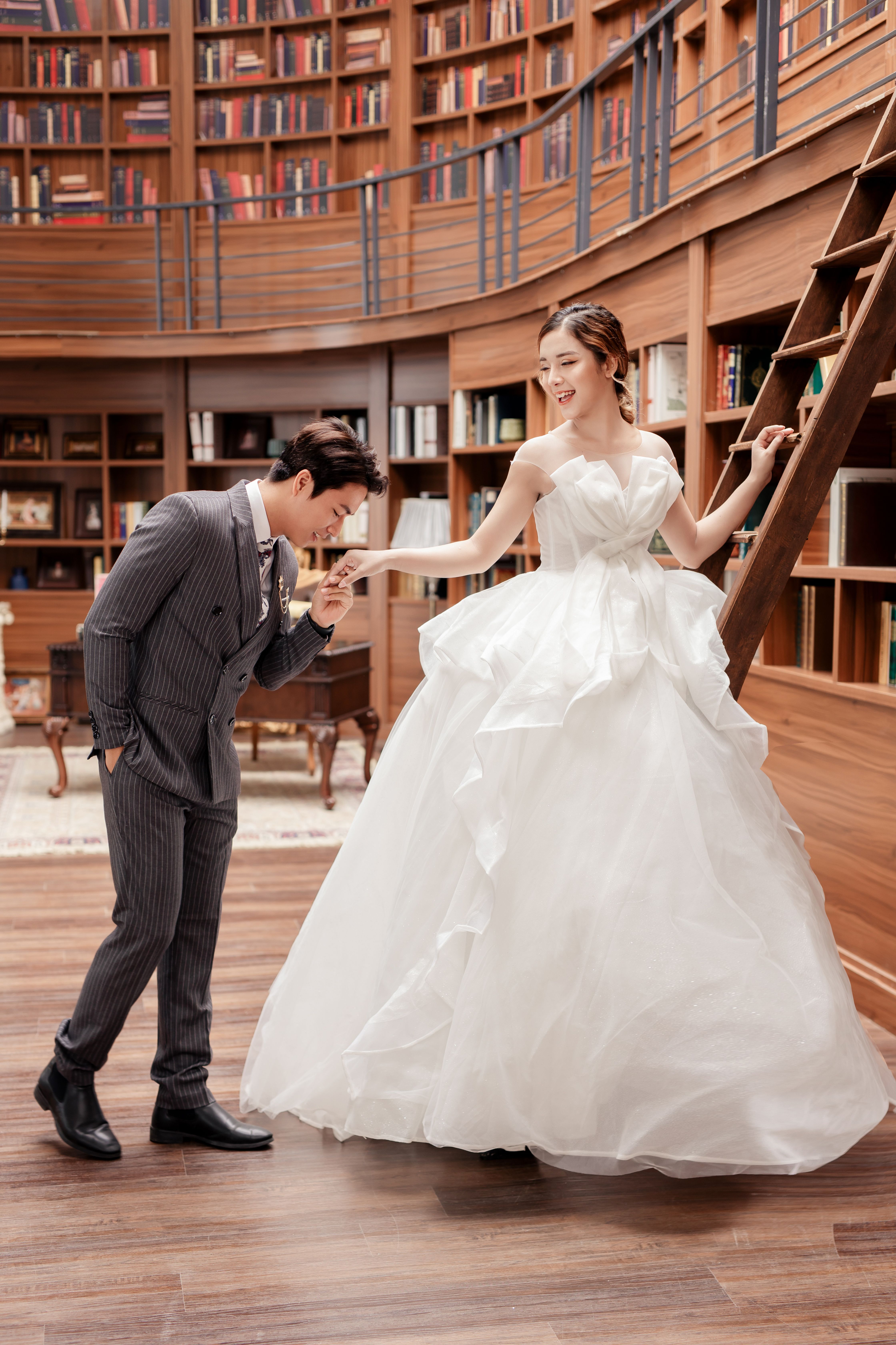Full length young Asian groom in chic tuxedo kissing happy brides hand while standing together in spacious library