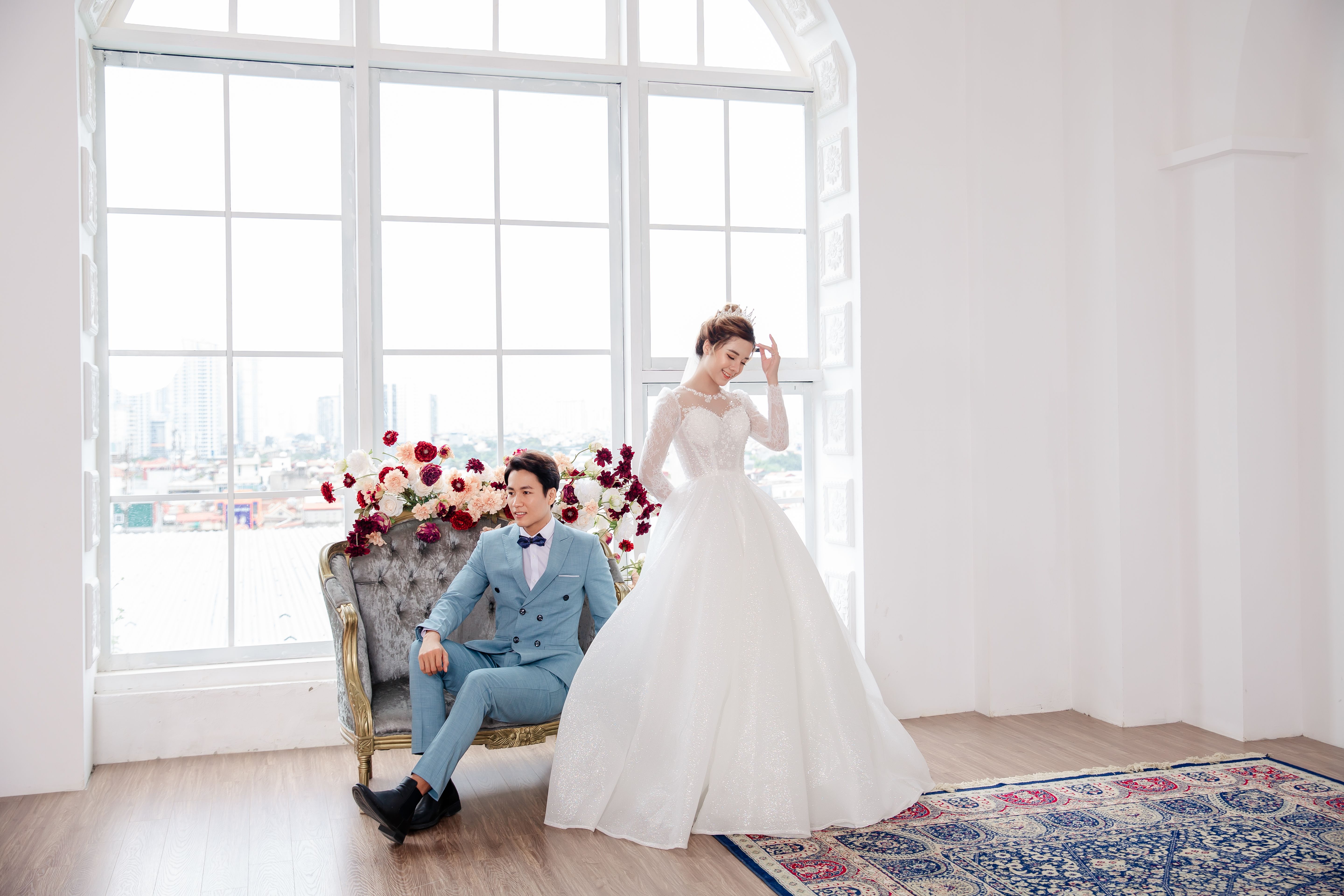 Full length happy Asian bride wearing chic  white dress standing near groom resting on classy sofa in light spacious studio