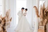 Side view full length cheerful young Asian bride touching grooms lips while hugging and looking at each other in fancy wedding studio