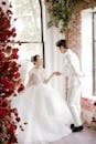 Full length cheerful newlywed Asian couple wearing elegant white wedding gowns holding hands and looking at each other with love