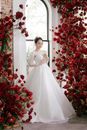 Full body serene young Asian bride wearing classy white dress standing in stylish wedding studio decorated with lush red flowers