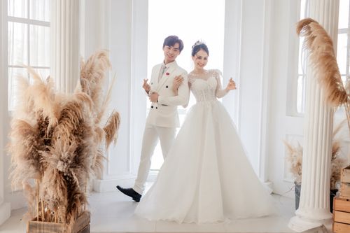 Free Full body smiling newlywed Asian couple in classy white gowns standing together in fancy wedding studio and looking at camera happily Stock Photo