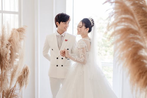 Romantic newlywed Asian couple wearing elegant wedding gowns standing in fancy wedding studio and touching hands