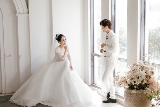 Side view of Asian elegant bride in white dress and finance in suit near window